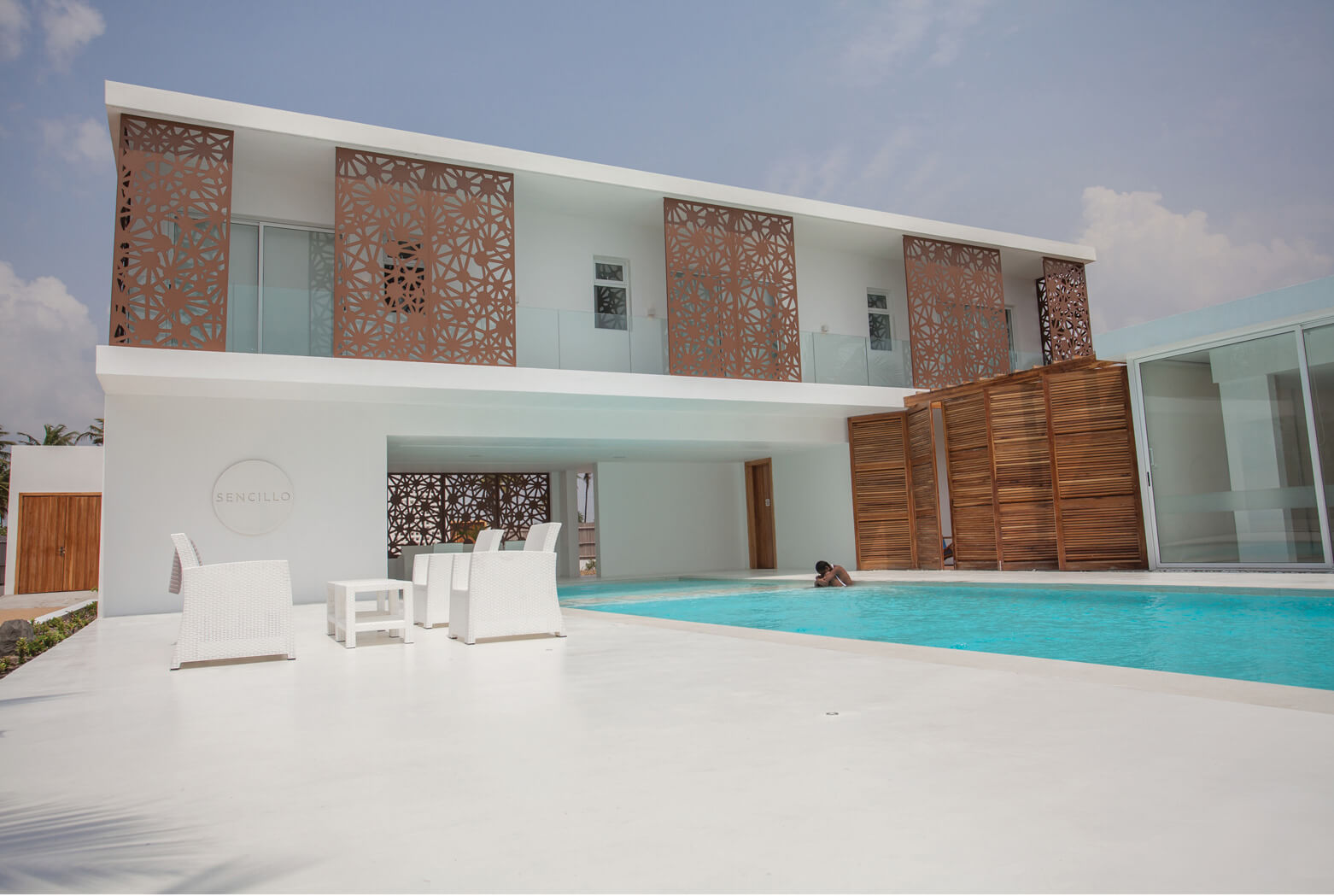 Sencillo Beach House Lagos showcases a modern architectural design by Oshinowo Studio, led by Tosin Oshinowo. The exterior features a clean, white façade, accented by unique, patterned lattice panels in a rust color. The property includes a large swimming pool with white lounge chairs around it, enhancing the luxury beachfront experience.
