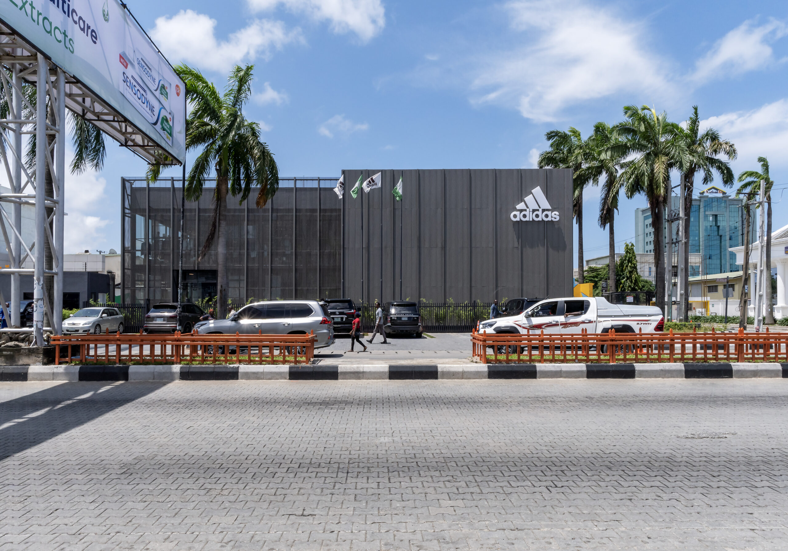 Daytime view of the ADIDAS Lagos flagship store, a two-story building with a modern, minimalist design featuring large glass windows and a metallic facade. Created by Oshinowo Studio led by Tosin Oshinowo, a leading architectural design studio in Lagos, the building stands out with its clean lines and the iconic ADIDAS logo mounted on the exterior wall. Palm trees flank the building, and a bustling street scene unfolds in the foreground, capturing the dynamic spirit of Lagos.