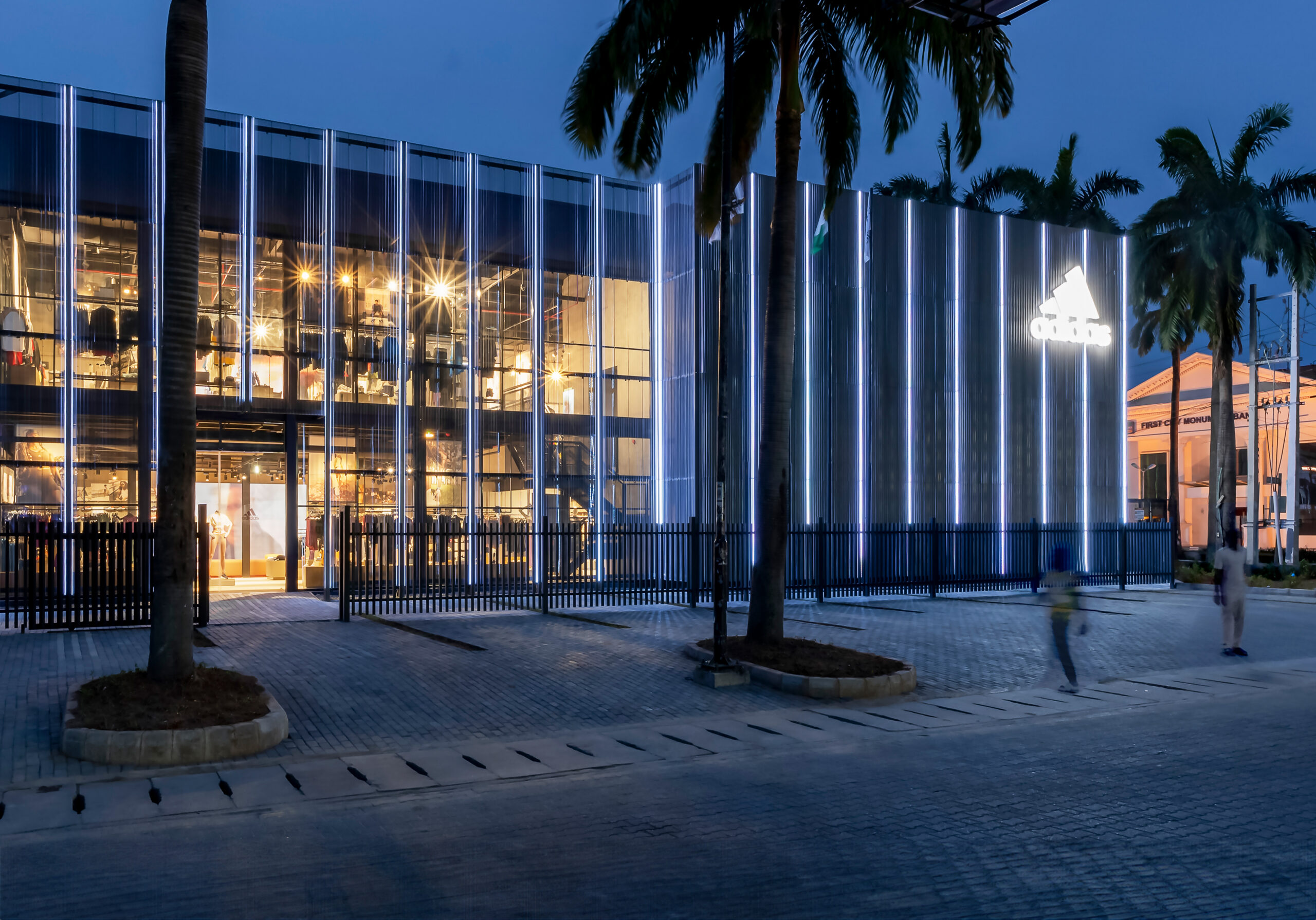 Evening view of the ADIDAS Lagos flagship store, a contemporary two-story building with a transparent glass facade that allows a peek into the brightly lit interior showcasing sportswear and accessories. The store's design, by the leading architectural design studio in Lagos, Oshinowo Studio, led by Tosin Oshinowo, features vertical illuminated stripes on the exterior walls creating a dynamic visual effect. Lush palm trees frame the structure, and a prominent ADIDAS logo glows above, casting a soft light on the surroundings. Pedestrians can be seen in motion, emphasizing the store's integration into the city's vibrant street life.