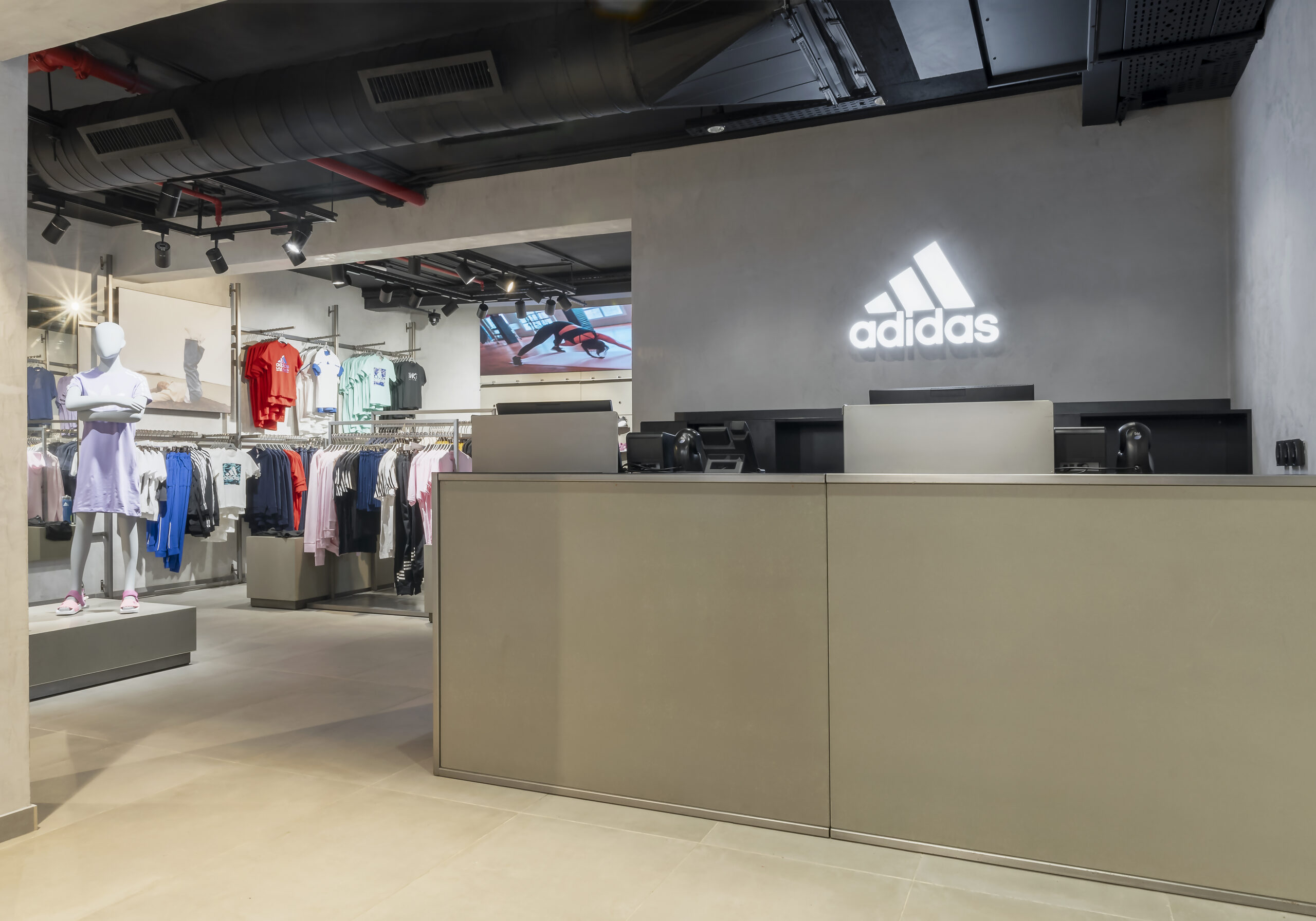 Interior of the ADIDAS Lagos flagship store, featuring a modern and industrial design by Oshinowo Studio, led by Tosin Oshinowo. The space is characterized by its exposed ceiling with visible ductwork, accentuated by sleek lighting fixtures. A large, backlit ADIDAS logo dominates the wall behind the checkout counter, which is styled in muted tones. Mannequins and clothing racks display the latest athletic wear, inviting customers to explore the merchandise in this cutting-edge retail environment.