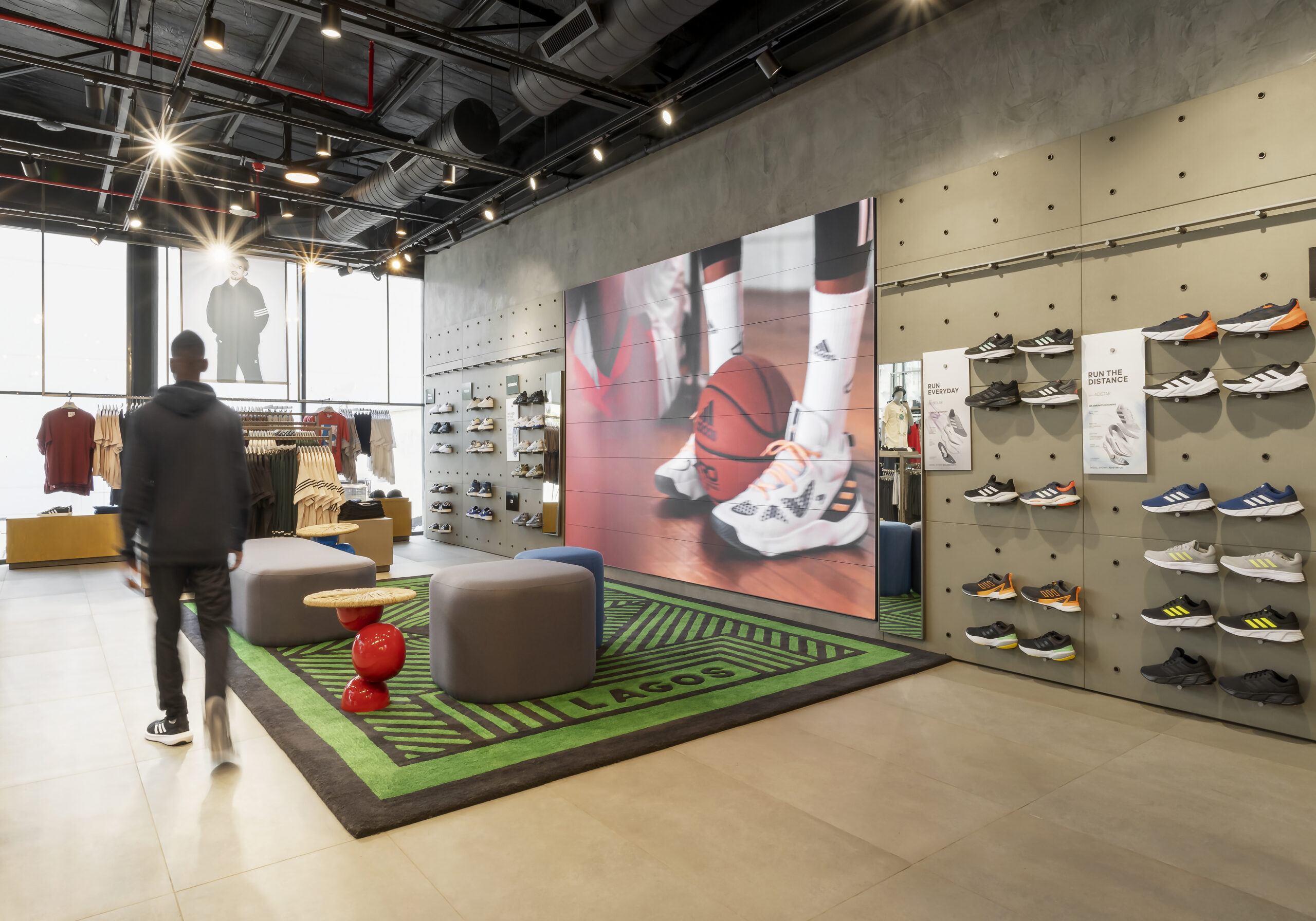 The interior of the ADIDAS Lagos flagship store, conceptualized by Oshinowo Studio, a leading architectural design studio in Lagos, led by Tosin Oshinowo, is captured in this image. The space features an urban-industrial aesthetic with exposed ceiling pipes, a concrete finish on walls, and a prominent display of athletic footwear on modular shelving. A large digital screen animates the space with sports imagery, while customers explore the variety of Adidas products, accentuated by strategic lighting that highlights the brand's fusion of performance and style.