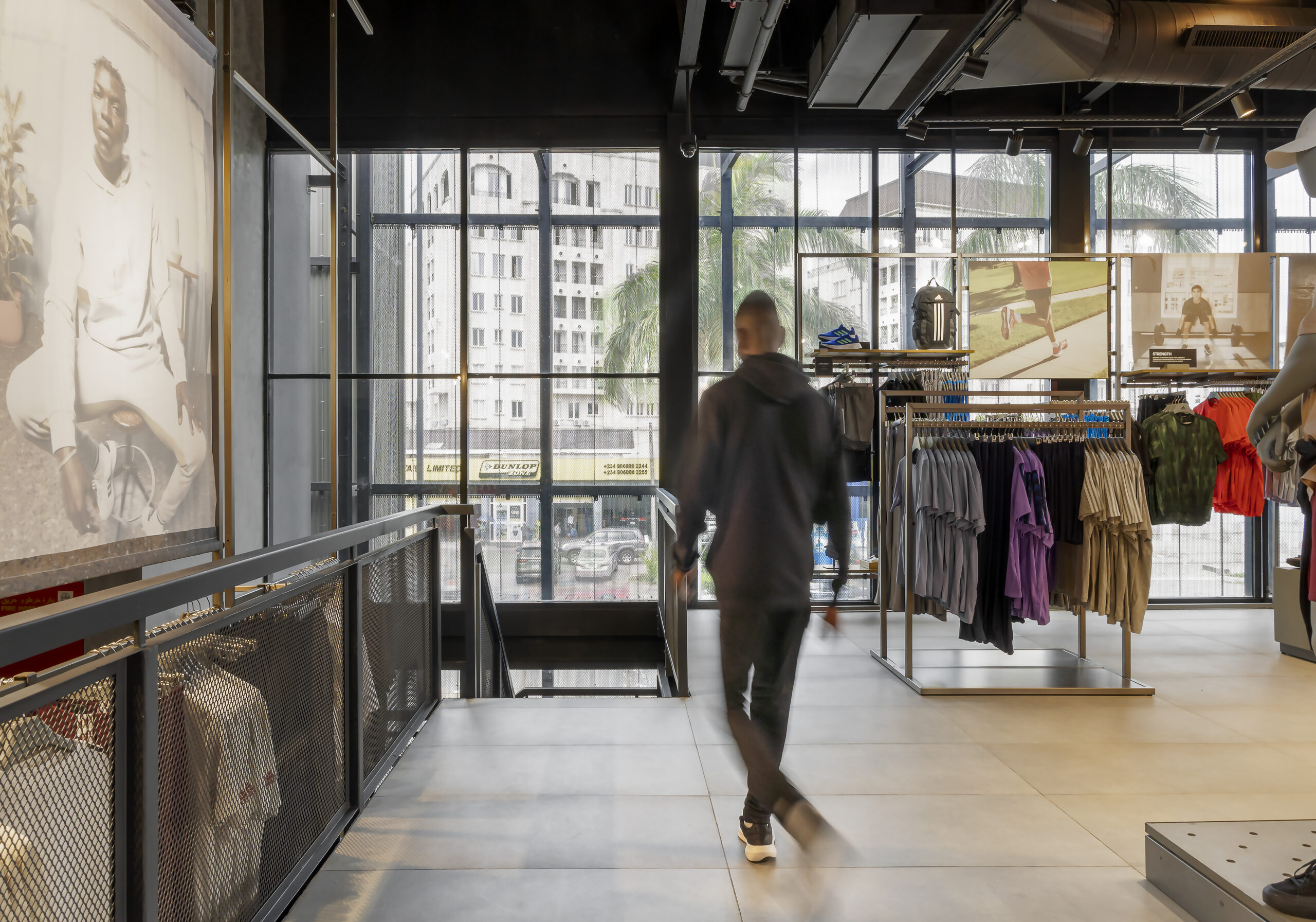 Inside the ADIDAS Lagos flagship store, designed by Oshinowo Studio, a leading architectural design studio in Lagos, led by Tosin Oshinowo, a customer is seen walking past a display. The store's interior features a blend of modern design elements such as a large lifestyle graphic, transparent glass windows offering a view of the city, and urban-industrial ceiling details. The array of neatly organized sportswear contributes to the store's sophisticated yet functional atmosphere.