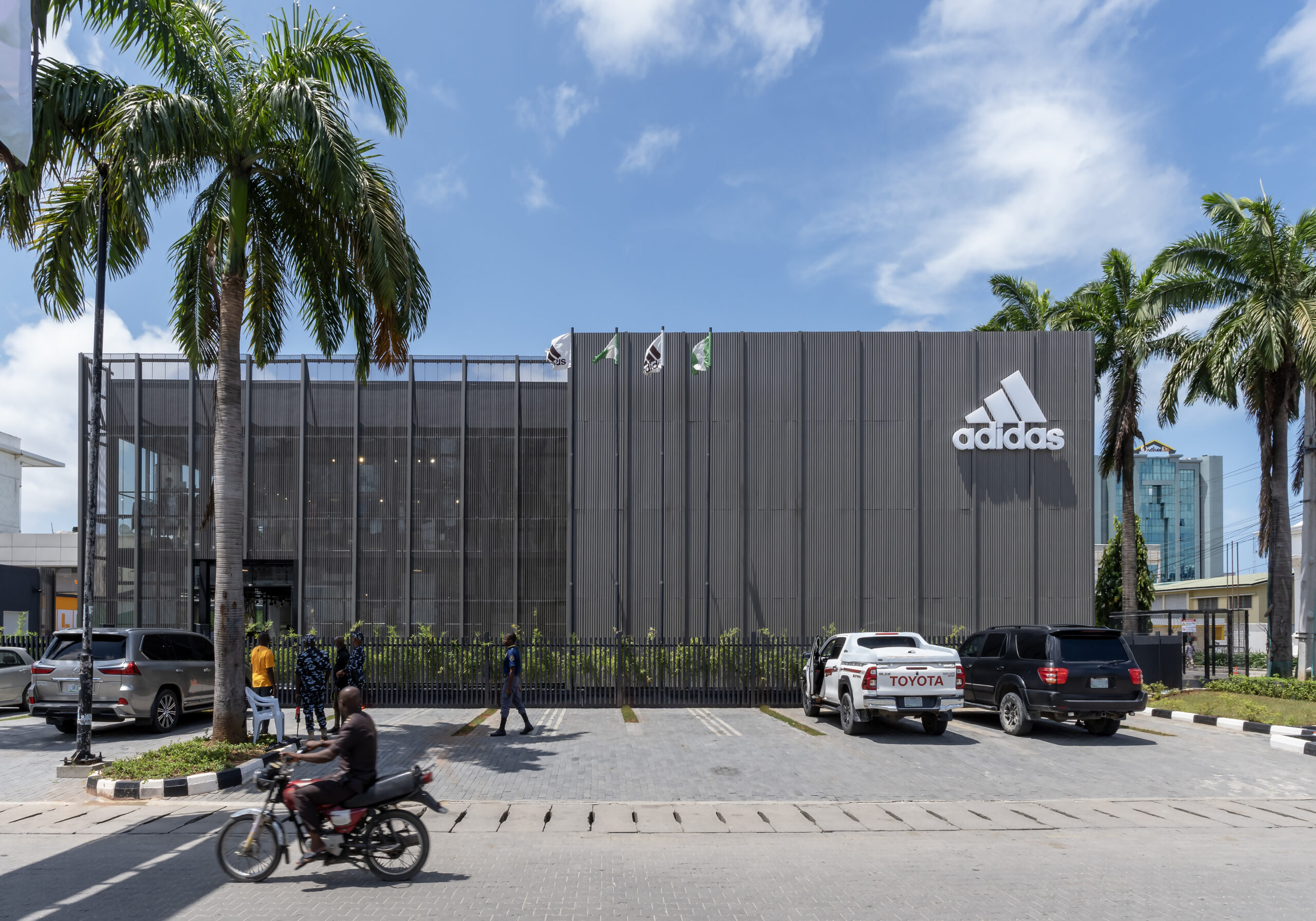 Exterior view of the ADIDAS Lagos flagship store during a bright day, showcasing the industrial-chic architecture by Oshinowo Studio, a leading architectural design studio in Lagos led by Tosin Oshinowo. The facade is a combination of gray metal panels and glass, with the iconic ADIDAS logo prominently affixed to the upper section of the building. Surrounded by lush palm trees and an active urban environment, including pedestrians and a motorcycle in motion, the store exudes a contemporary vibe in the heart of Lagos.