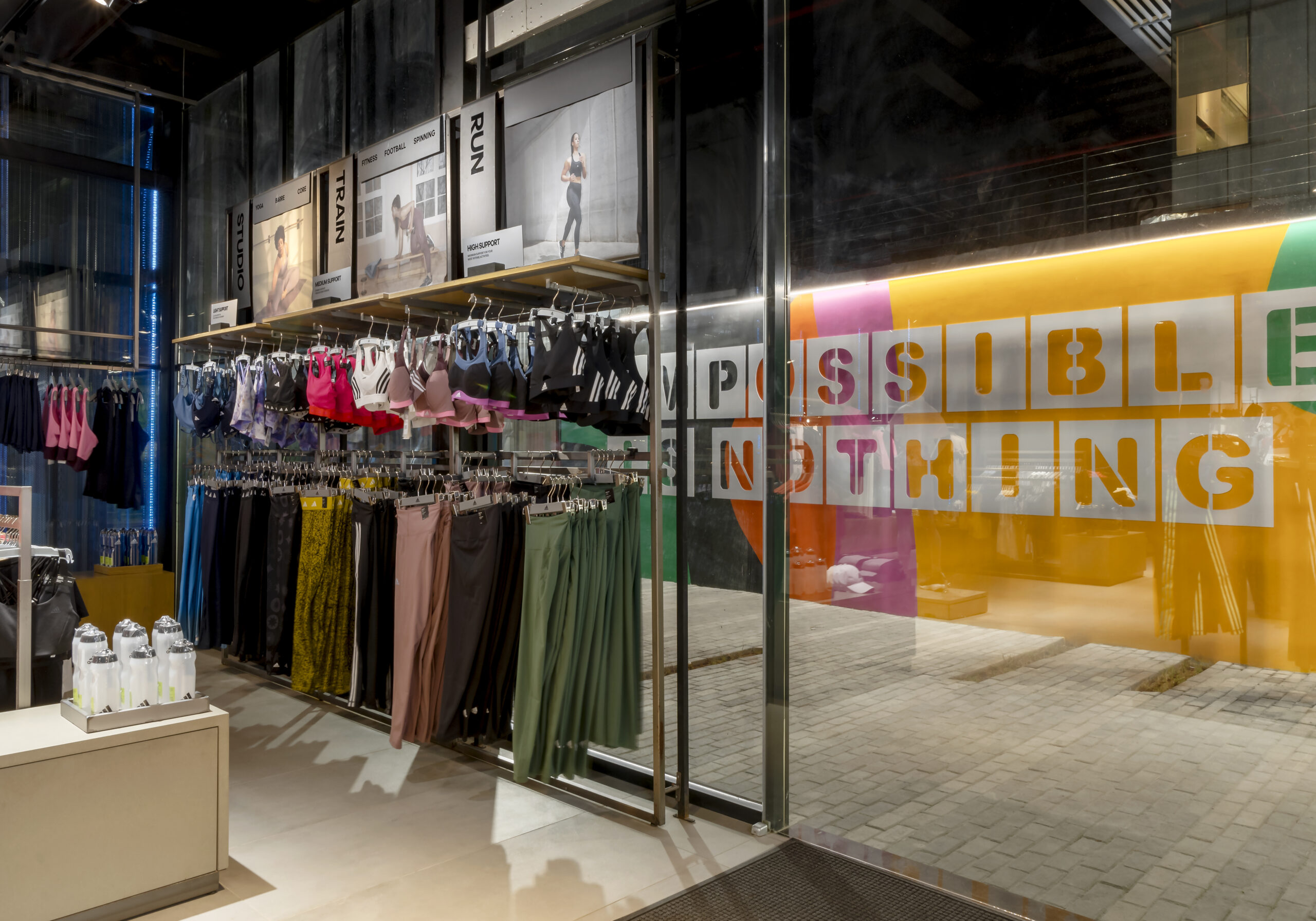 The image captures a sleek and modern corner of the ADIDAS flagship store, a project touched by the creative flair of Oshinowo Studio, a leading architectural design studio in Lagos. The space is meticulously organized, with sports bras in a spectrum of colors hanging above complementary fitness apparel, and an inspirational “IMPOSSIBLE IS NOTHING” graphic adorning a transparent divider, encapsulating the brand's empowering philosophy within the refined and elegant retail environment.
