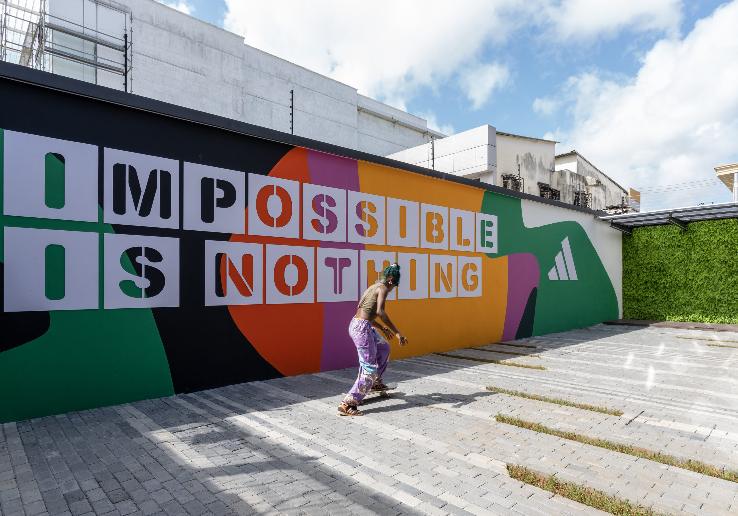 A person walking past a large, colorful mural on the exterior wall of the ADIDAS flagship store, designed by a leading architectural design studio in Lagos. The vibrant artwork features overlapping geometric shapes and bold text that reads "IMPOSSIBLE IS NOTHING". The modern building facade is visible in the background, along with some greenery.