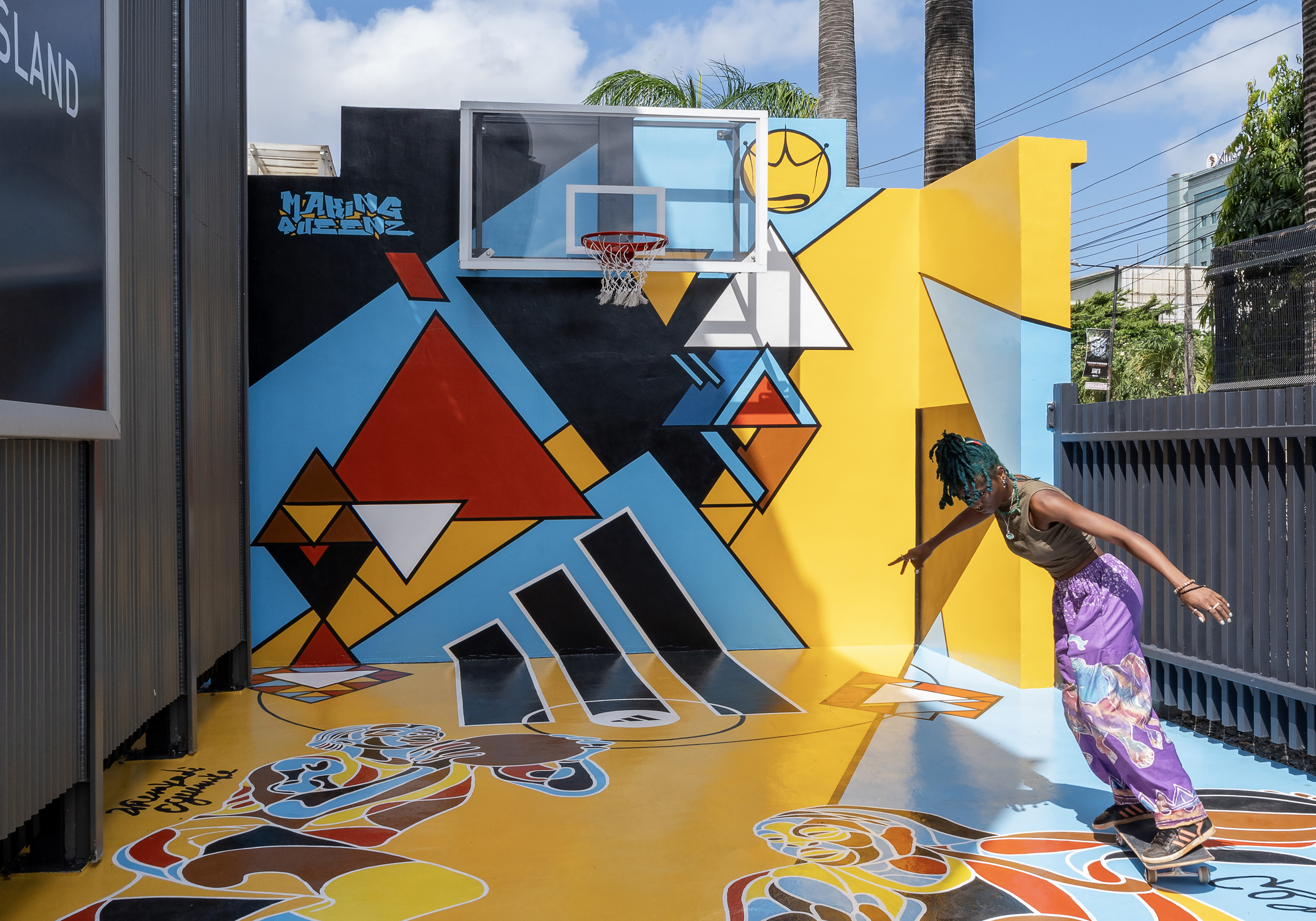 The outdoor basketball court art installation. The colorful, geometric mural covers the court surface, featuring abstract shapes in blue, red, yellow and brown tones. A person in patterned pants is shown in motion, seemingly jumping or dancing in front of the vibrant art piece designed by a leading architectural design studio in Lagos. Palm trees and the corner of a modern building are visible in the background.