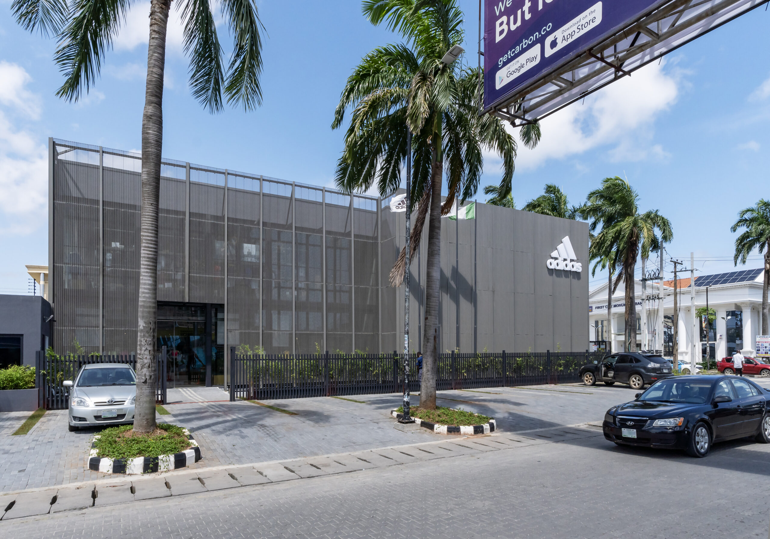 Exterior view of the ADIDAS Lagos flagship store during a bright day, showcasing the industrial-chic architecture by Oshinowo Studio, a leading architectural design studio in Lagos led by Tosin Oshinowo. The facade is a combination of gray metal panels and glass, with the iconic ADIDAS logo prominently affixed to the upper section of the building. Surrounded by lush palm trees and an active urban environment, including pedestrians and a motorcycle in motion, the store exudes a contemporary vibe in the heart of Lagos.