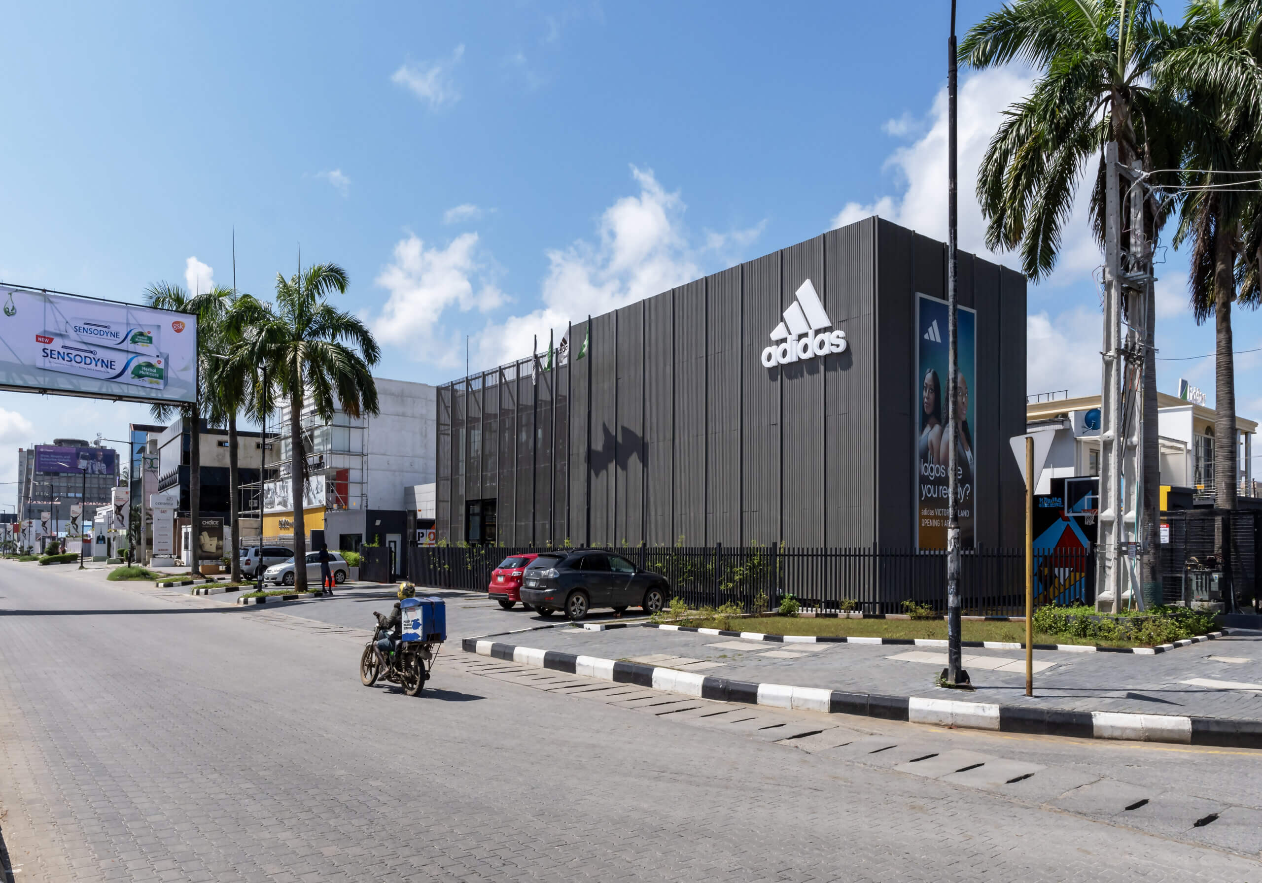 View of the ADIDAS Lagos flagship store, a robust structure with a metallic grey facade designed by Oshinowo Studio, a leading architectural design studio in Lagos led by Tosin Oshinowo. The store's frontage is adorned with the Adidas logo, flanked by Nigerian flags. Palm trees add a tropical touch to the urban setting. A blue sky looms above the busy street, where a motorbike and cars pass by, illustrating the lively atmosphere around this iconic retail landmark.