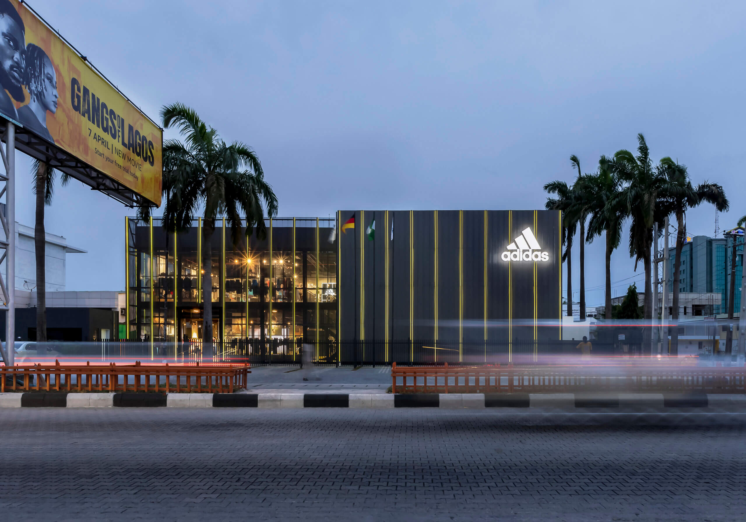 Twilight view of the ADIDAS Lagos flagship store, an architectural project by Oshinowo Studio led by Tosin Oshinowo, notable for its reflective glass exterior and vertical metal siding. The store's illuminated interior spills warm light into the early evening, with the ADIDAS logo brightly lit against the dusk sky. Palm trees and a billboard for a local movie add to the urban landscape, while the long exposure captures the streaks of headlights from the bustling traffic, conveying a sense of motion and the vibrant energy of Lagos.