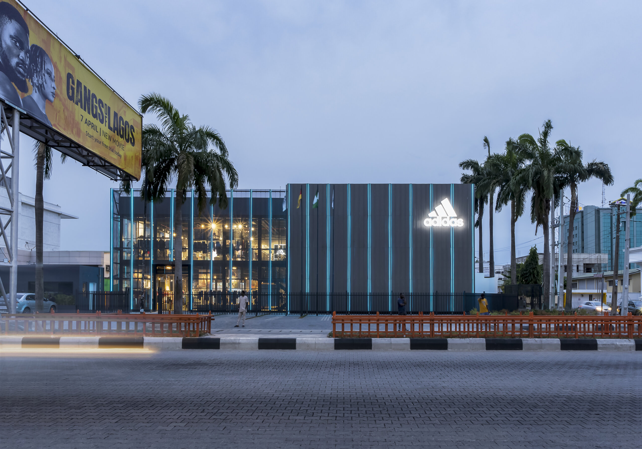 Twilight view of the ADIDAS Lagos flagship store, an architectural project by Oshinowo Studio, led by Tosin Oshinowo, notable for its reflective glass exterior and vertical metal siding. The store's illuminated interior spills warm light into the early evening, with the ADIDAS logo brightly lit against the dusk sky. Palm trees and a billboard for a local movie add to the urban landscape, while the long exposure captures the streaks of headlights from the bustling traffic, conveying a sense of motion and the vibrant energy of Lagos.