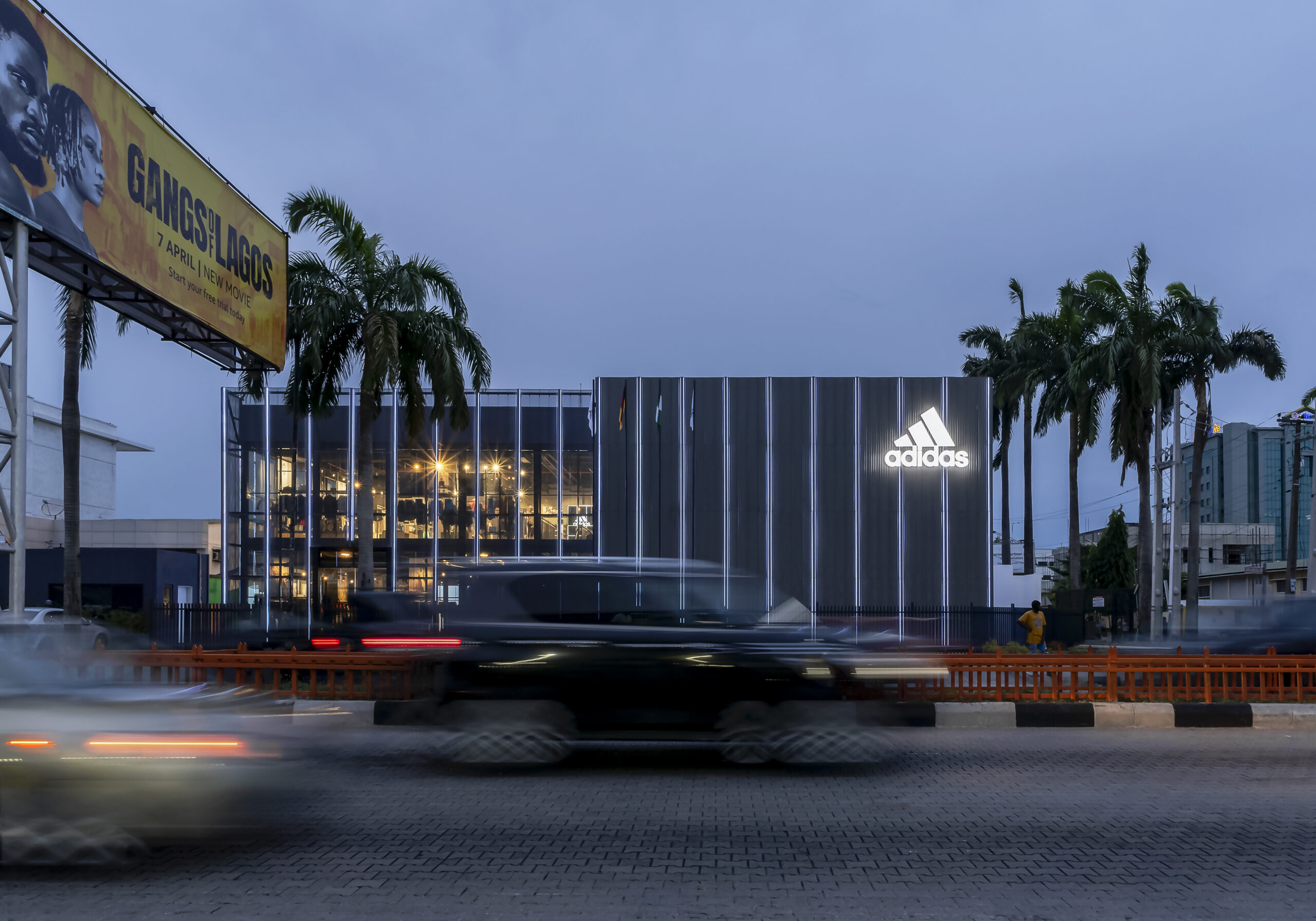 Twilight view of the ADIDAS Lagos flagship store, an architectural project by Oshinowo Studio, led by Tosin Oshinowo, notable for its reflective glass exterior and vertical metal siding. The store's illuminated interior spills warm light into the early evening, with the ADIDAS logo brightly lit against the dusk sky. Palm trees and a billboard for a local movie add to the urban landscape, while the long exposure captures the streaks of headlights from the bustling traffic, conveying a sense of motion and the vibrant energy of Lagos.