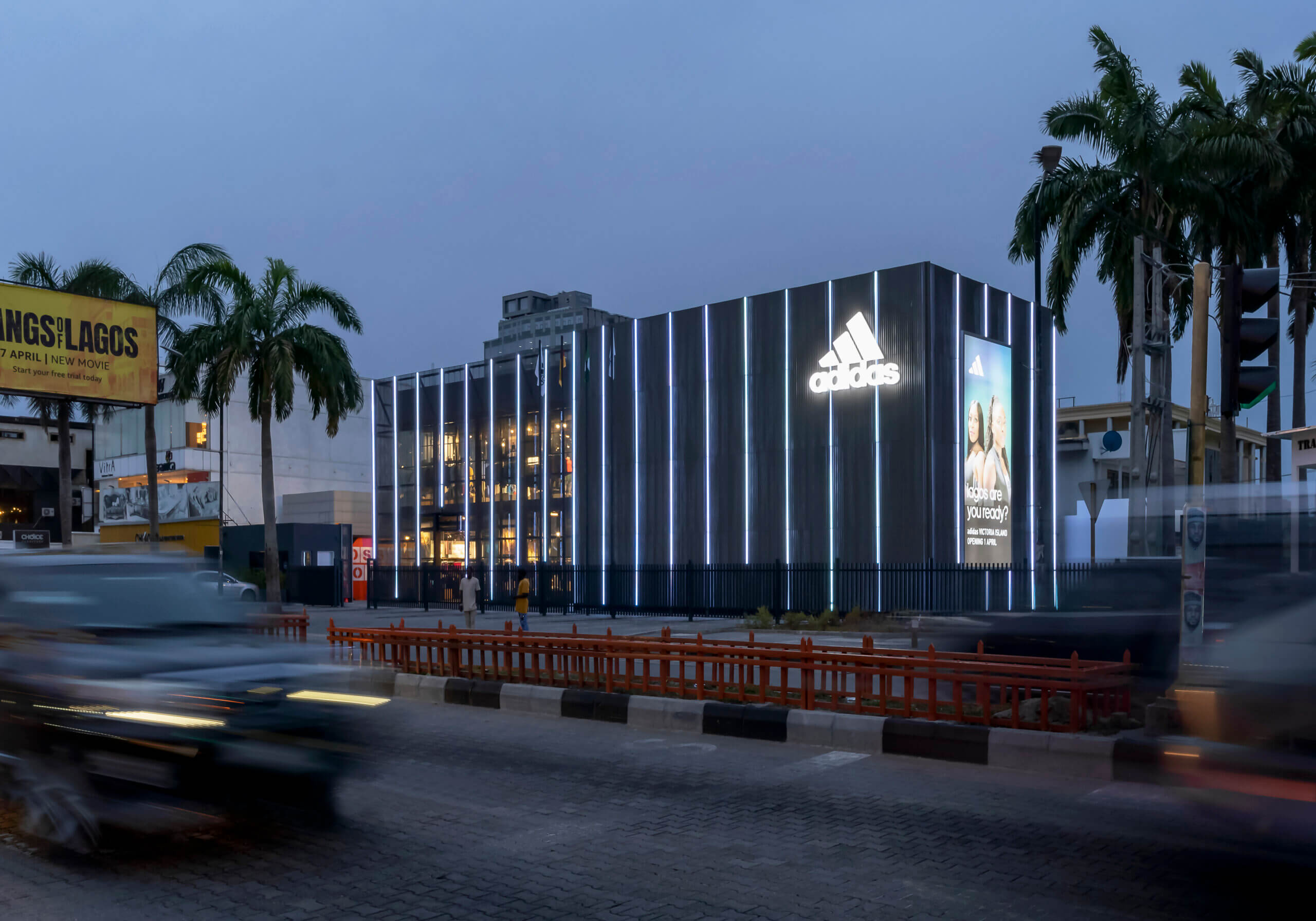 Twilight view of the ADIDAS flagship store, an architectural project by Oshinowo Studio, led by Tosin Oshinowo, notable for its reflective glass exterior and vertical metal siding. The store's illuminated interior spills warm light into the early evening, with the ADIDAS logo brightly lit against the dusk sky. Palm trees and a billboard for a local movie add to the urban landscape, while the long exposure captures the streaks of headlights from the bustling traffic, conveying a sense of motion and the vibrant energy of Lagos.