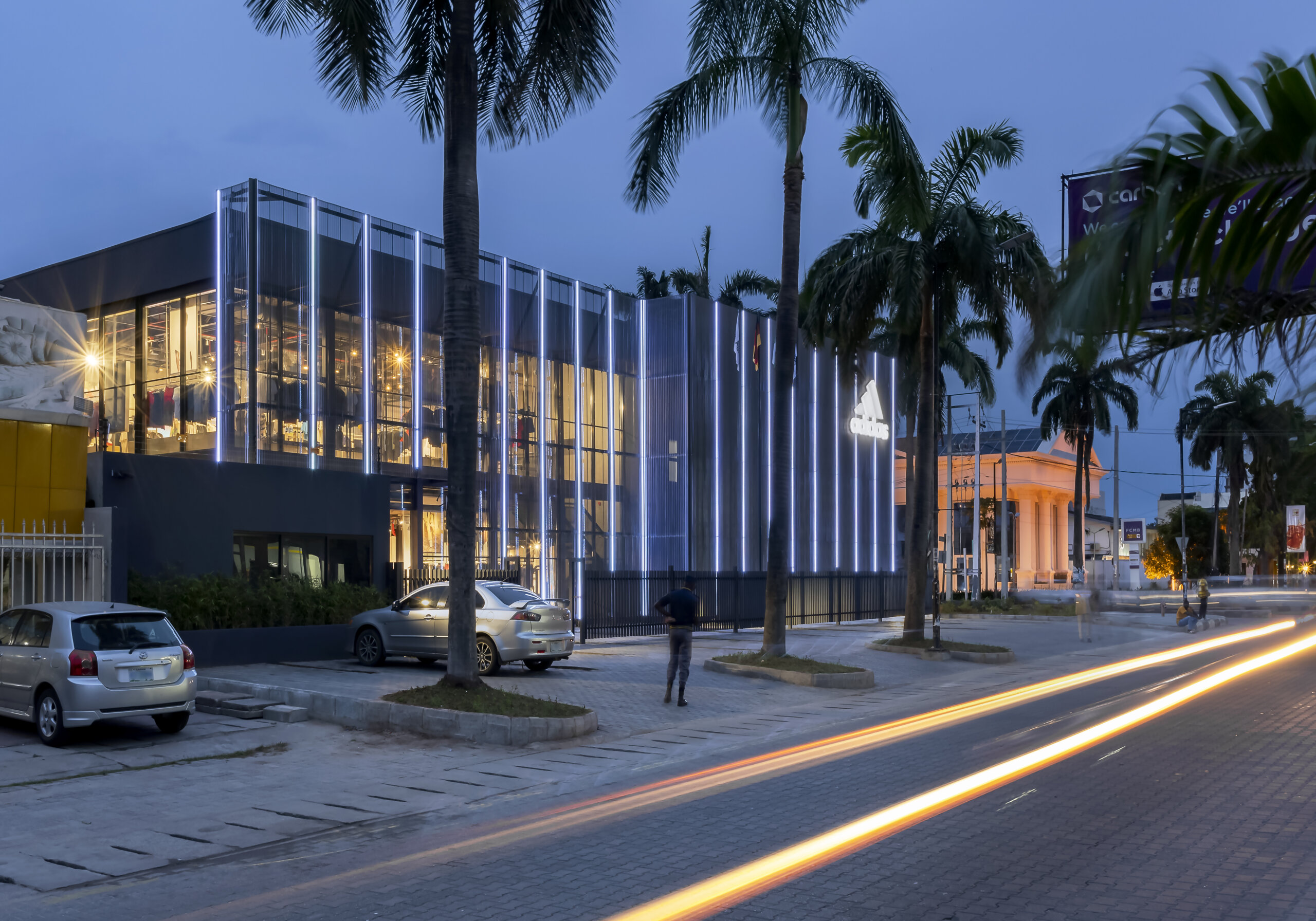 Twilight view of the ADIDAS Lagos flagship store, an architectural project by Oshinowo Studio, notable for its reflective glass exterior and vertical metal siding. The store's illuminated interior spills warm light into the early evening, with the ADIDAS logo brightly lit against the dusk sky. Palm trees and a billboard for a local movie add to the urban landscape, while the long exposure captures the streaks of headlights from the bustling traffic, conveying a sense of motion and the vibrant energy of Lagos. This project was led by Tosin Oshinowo.